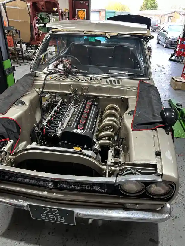 A front-facing photo of a beige classic Nissan Skyline GT-R with its hood open, revealing a custom engine with red valve…