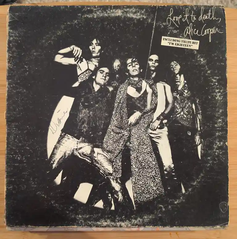 A black-and-white, worn album cover of Alice Cooper's 'Love It to Death' featuring the band in theatrical, dark attire. The…