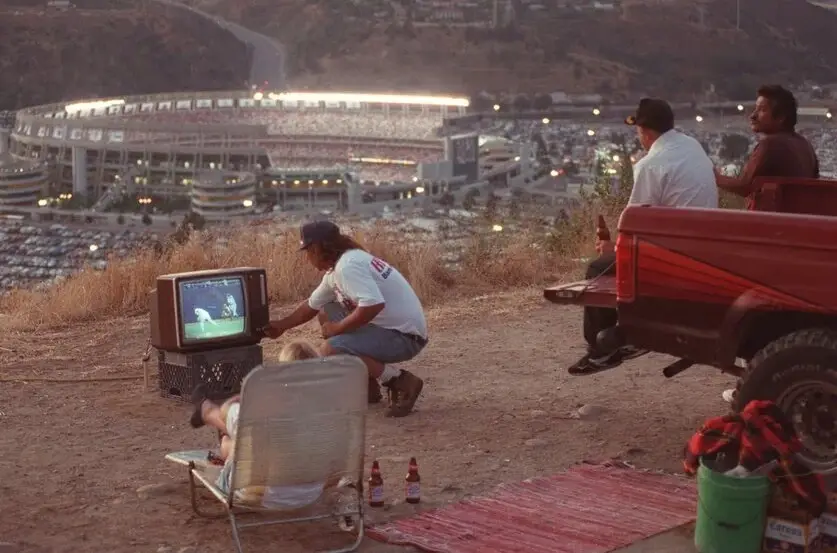 A group of people are gathered outdoors on a hillside at dusk, watching a baseball game on a small portable television. One…