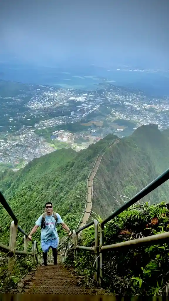 A man stands at the top of a steep, narrow mountain staircase with a vast cityscape and ocean visible far below. He holds…