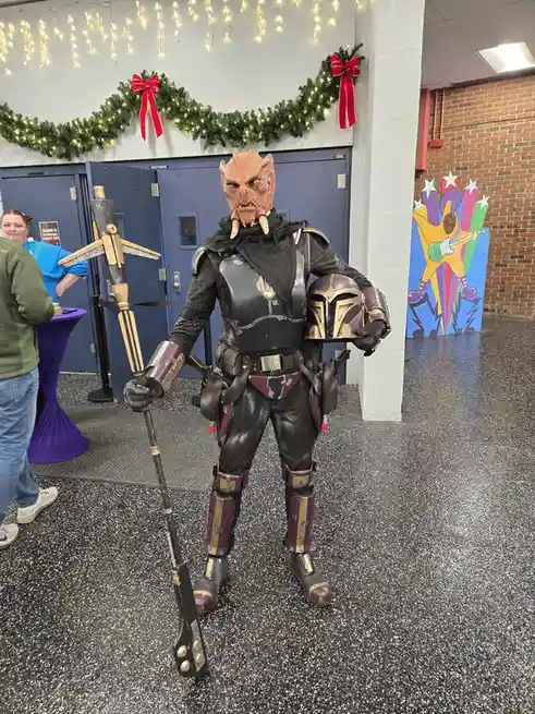A person in a detailed Mandalorian cosplayer costume, holding a blaster rifle and a Mandalorian helmet, stands in a hallway…
