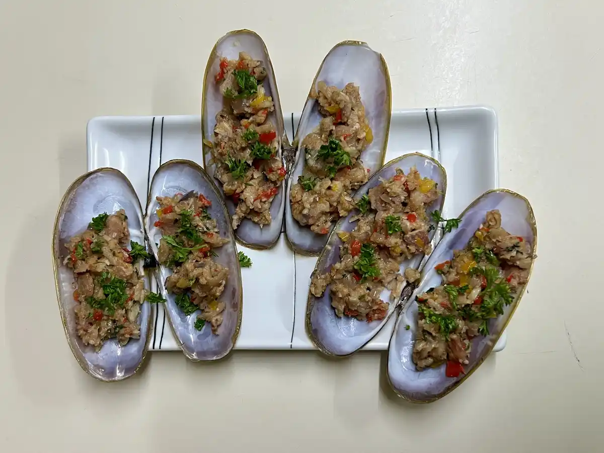 A white rectangular plate holds six open razor clams, each filled with a savory mixture of chopped ingredients including red…