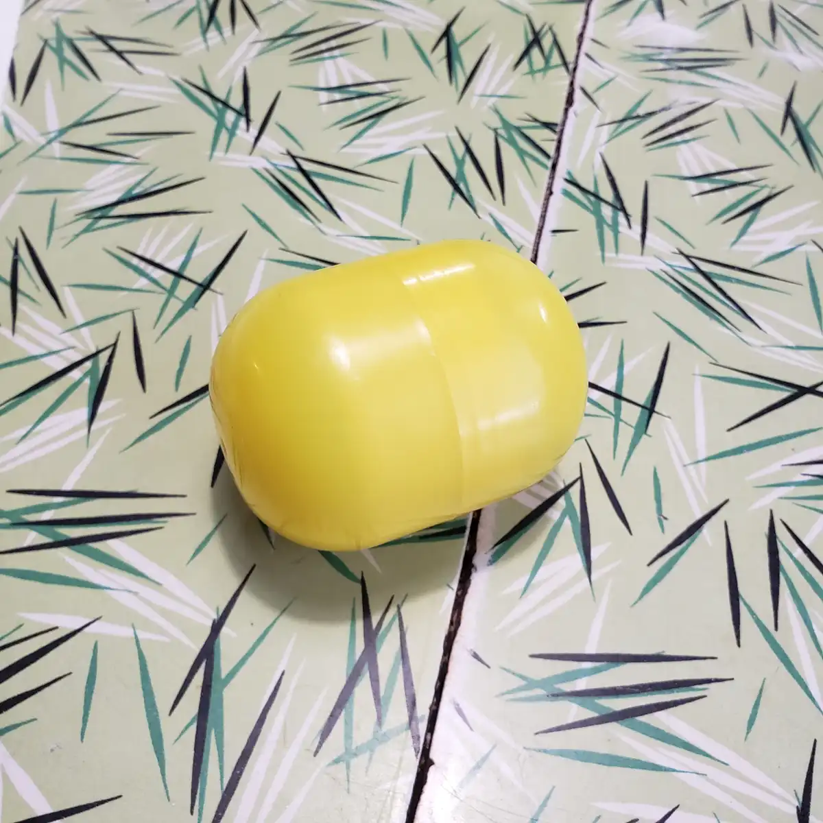 A bright yellow, smooth, oval-shaped plastic object resembling a potato sits on a light green surface with a pattern of…