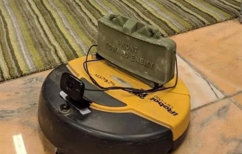 A yellow and black iRobot Roomba vacuum cleaner has a green military-style ammo box mounted on top, with the text 'FRONT…