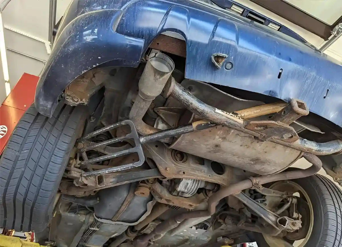 A low-angle shot of the underside of a blue car on a lift in a garage, showing rusted undercarriage, exhaust pipes, and…