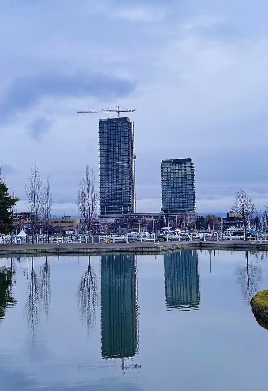 A calm waterfront scene with two tall glass skyscrapers under construction, one with a crane on top, reflected perfectly in…