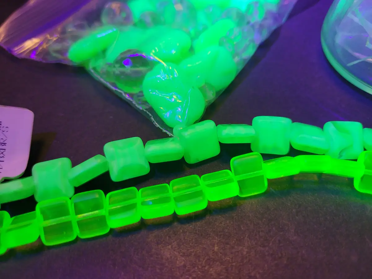 A close-up photo shows two strands of bright green, glowing plastic beads on a dark surface, with a bag of similar glowing…