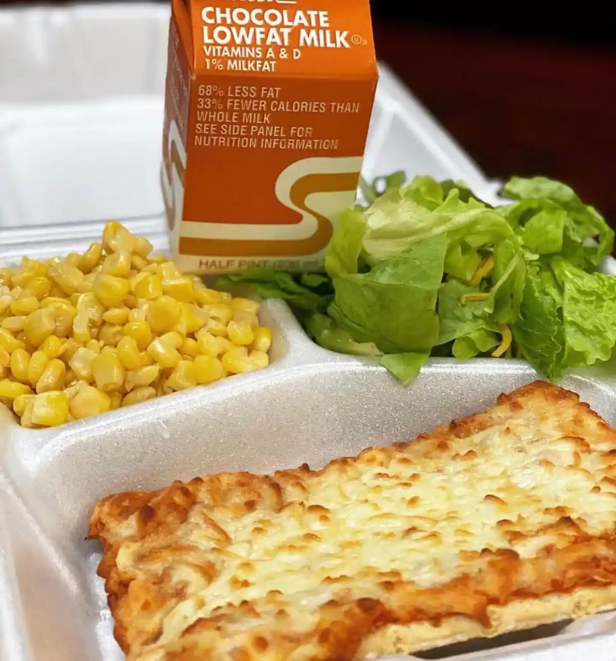 A close-up of a school lunch tray containing a cheesy pizza slice, a side of corn, and a side of lettuce, with a carton of…