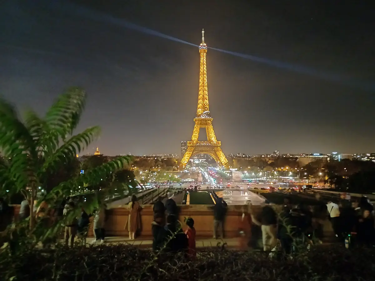 A nighttime photo of the Eiffel Tower in Paris, illuminated and casting beams into the sky, with blurred crowds of people in…