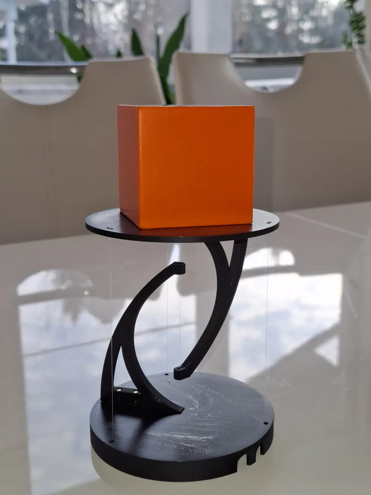 A solid orange cube sits atop a black, ornate, three-legged stand on a white table, with chairs and plants visible in the…