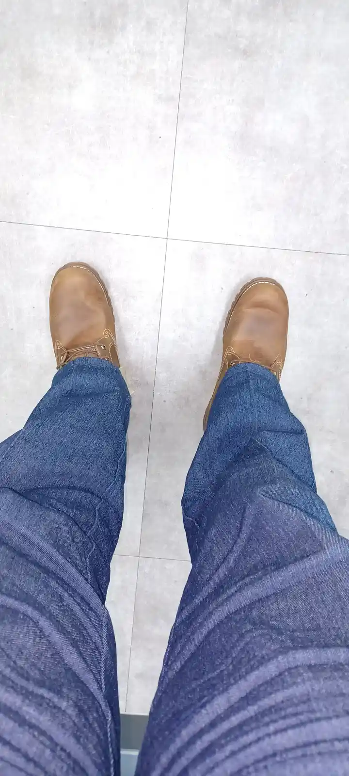 A top-down view shows a person's legs in blue jeans and tan leather lace-up boots standing on a light gray tiled floor. The…