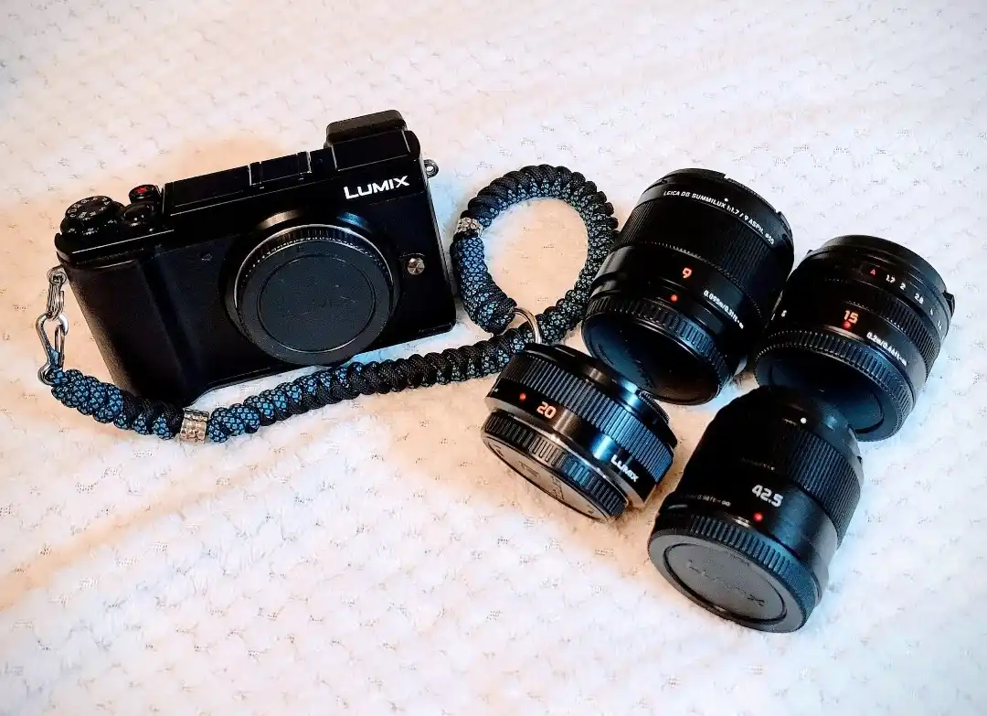 A black Lumix camera with a braided strap lies on a textured white surface, surrounded by four lenses labeled 9, 15, 20, and…