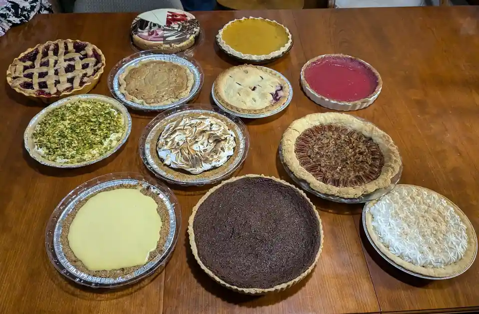 A wooden table is covered with a dozen different pies in various pans, including lattice-top berry, chocolate, pecan…