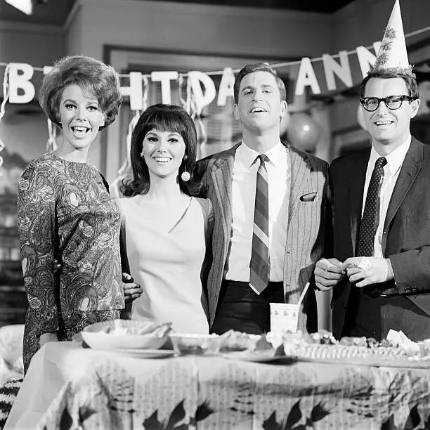 A black-and-white photo of four smiling people at a 1960s birthday party, with a banner reading 'BIRTHDAY ANN' behind them…