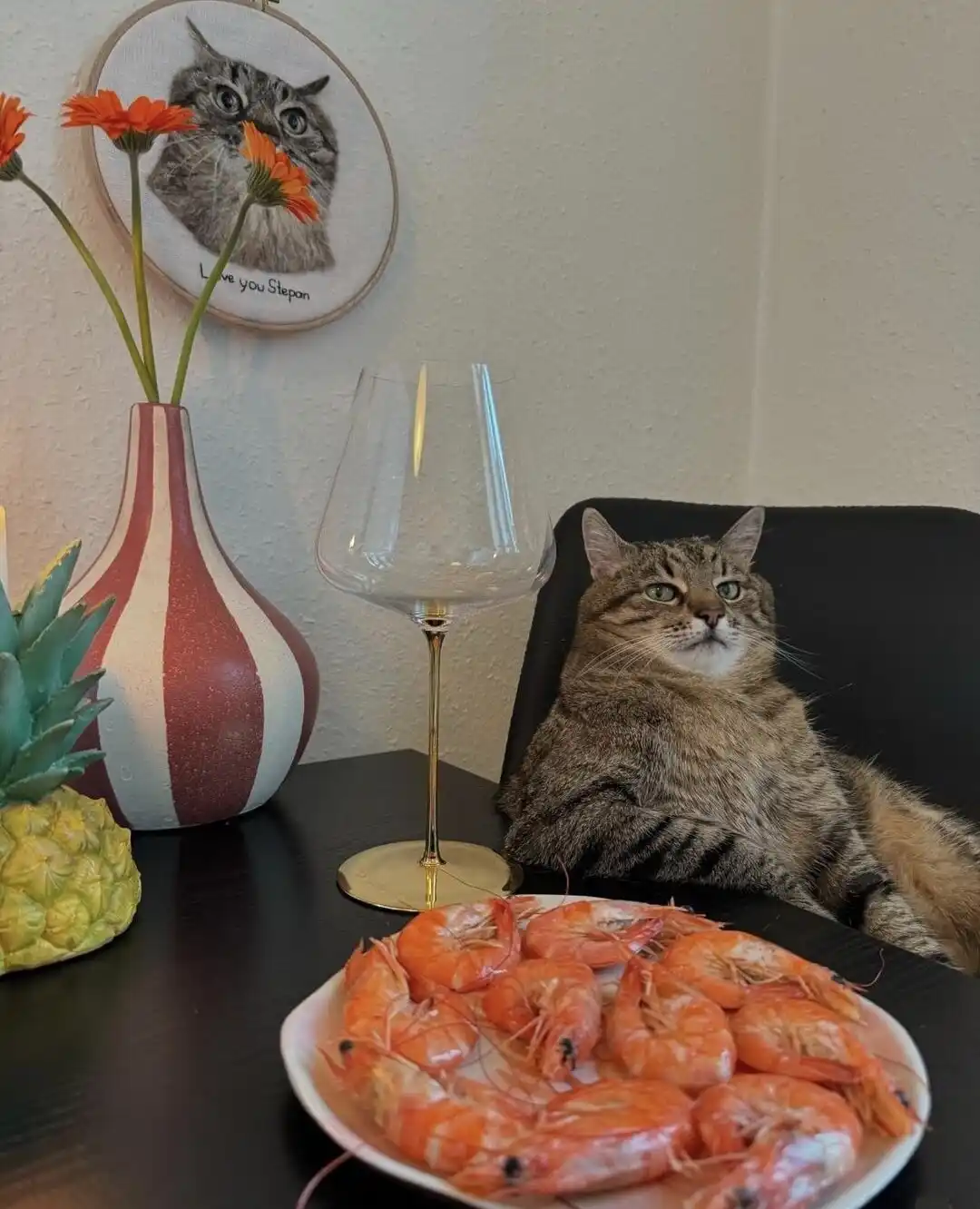 A tabby cat sits upright in a black chair beside a table with a plate of cooked shrimp, an empty wine glass, a striped vase…