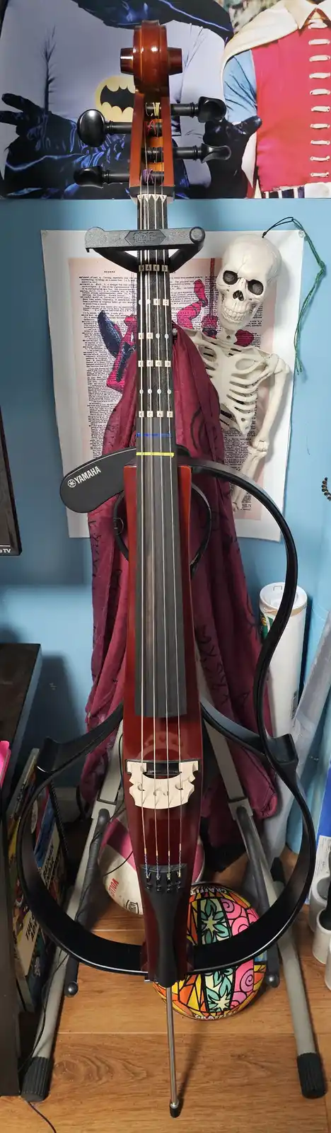 A Yamaha cello with a black circular stand is positioned upright in a room, with a skeleton decoration visible behind it…
