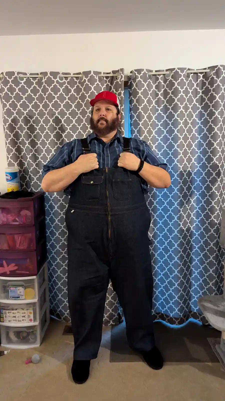 A man with a beard and red baseball cap stands indoors wearing denim overalls over a plaid shirt, gripping the suspenders…