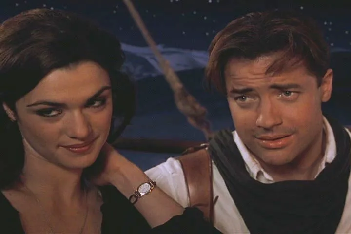 A still from a movie shows a woman with dark hair and a watch smiling smugly while resting her hand on a man's shoulder; the…