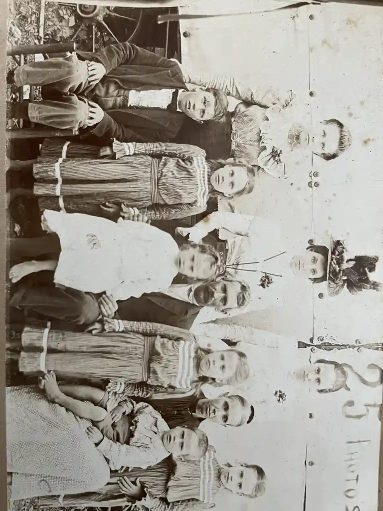 A sepia-toned, vertically oriented vintage family portrait shows a group of adults and children posing formally outdoors…