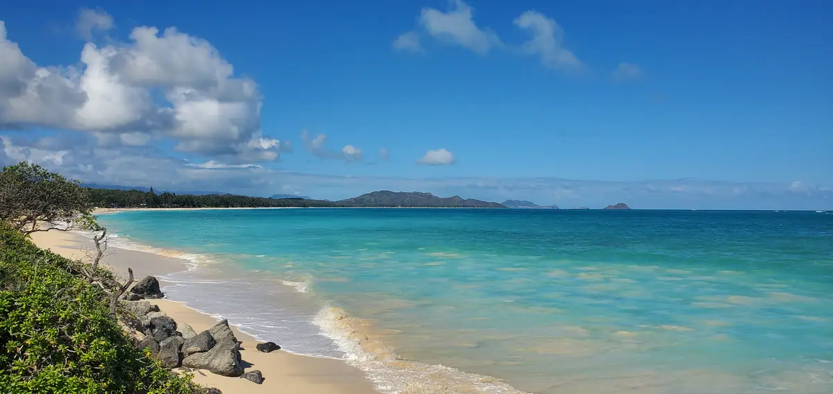 A scenic tropical beach with turquoise water, white sand, and distant mountains under a blue sky with fluffy clouds. The…