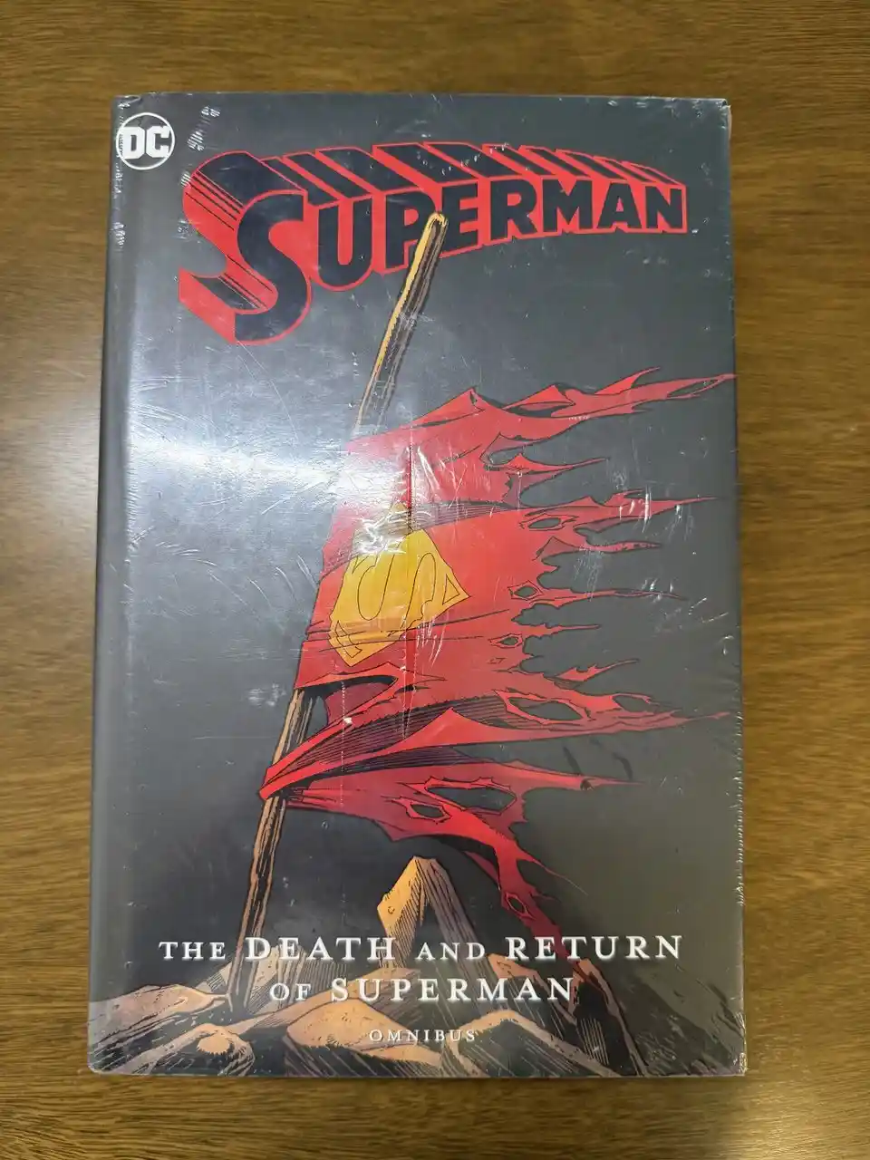 A DC Comics hardcover omnibus titled 'The Death and Return of Superman' is shown wrapped in plastic, resting on a wooden…