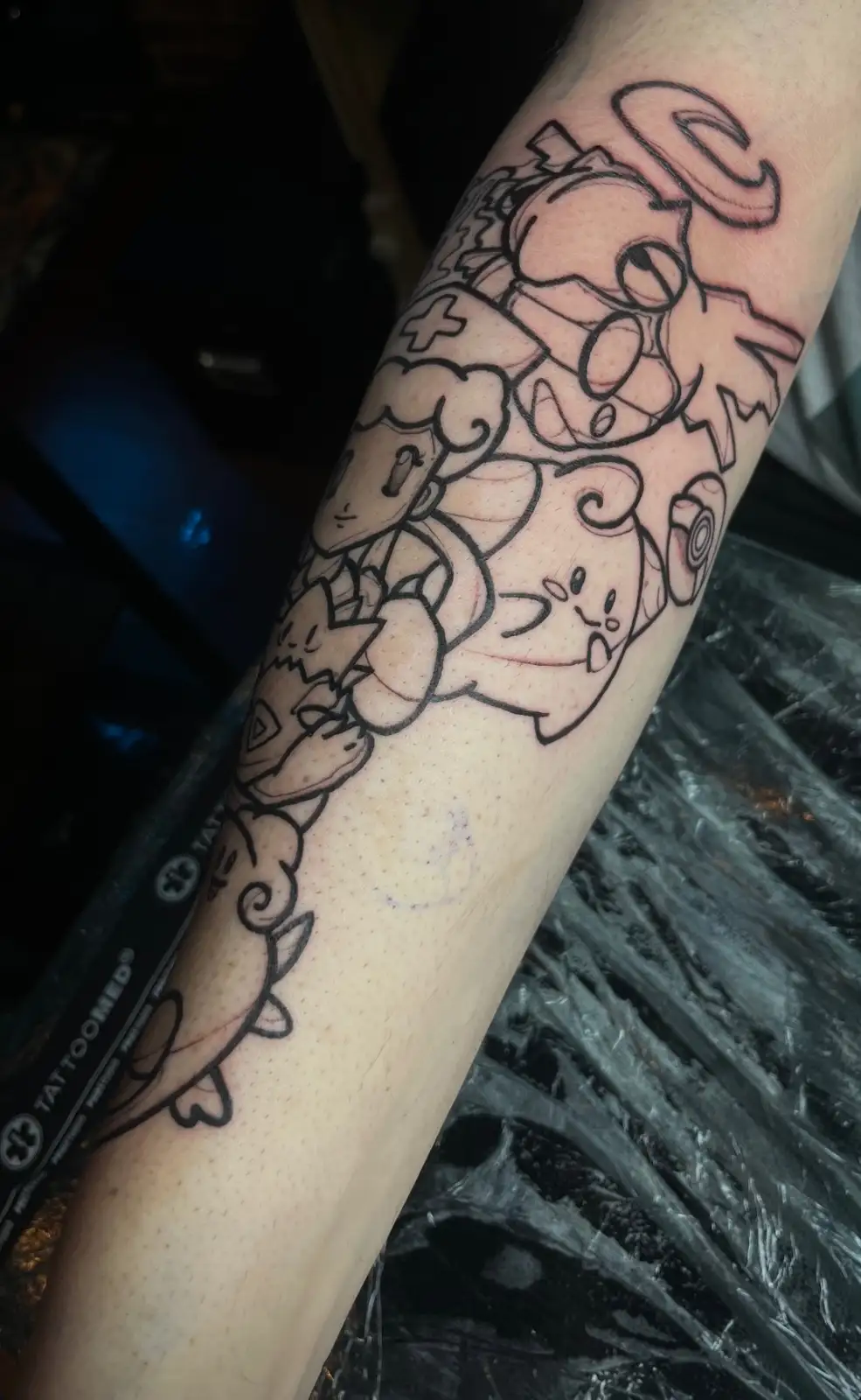 A freshly inked tattoo on a person's forearm features a dense, black-lined design of multiple Pokémon characters, including…