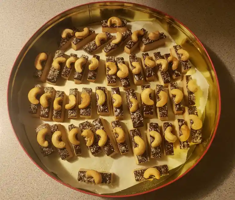 A golden tin contains neatly arranged rectangular chocolate-covered treats, each topped with a single cashew nut. The text…