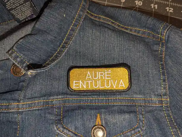 A close-up of a blue denim jacket with yellow stitching, showing a rectangular yellow patch with white embroidered text…