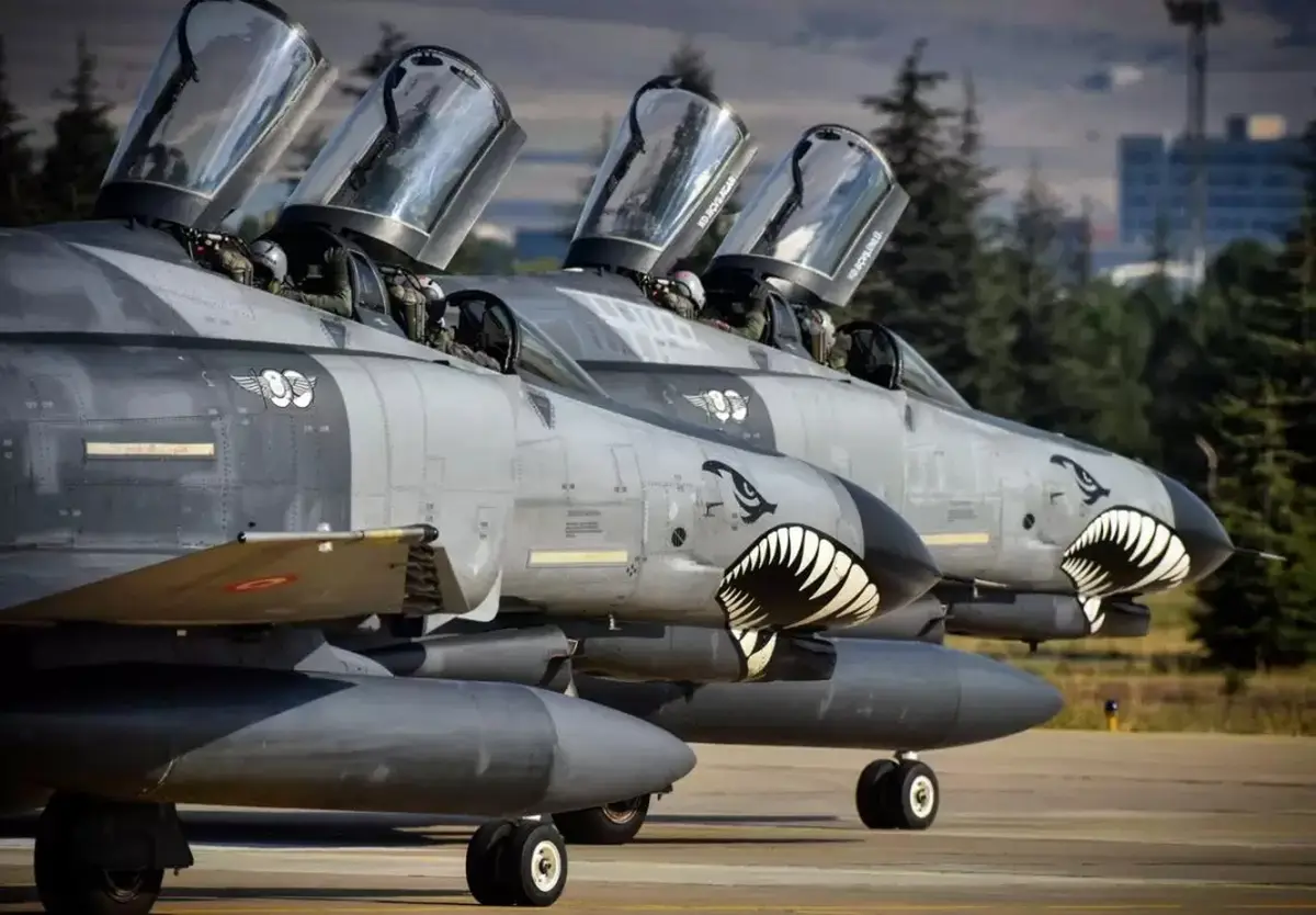 A row of grey fighter jets with their canopies open and pilots visible, each featuring a menacing shark mouth design on the…