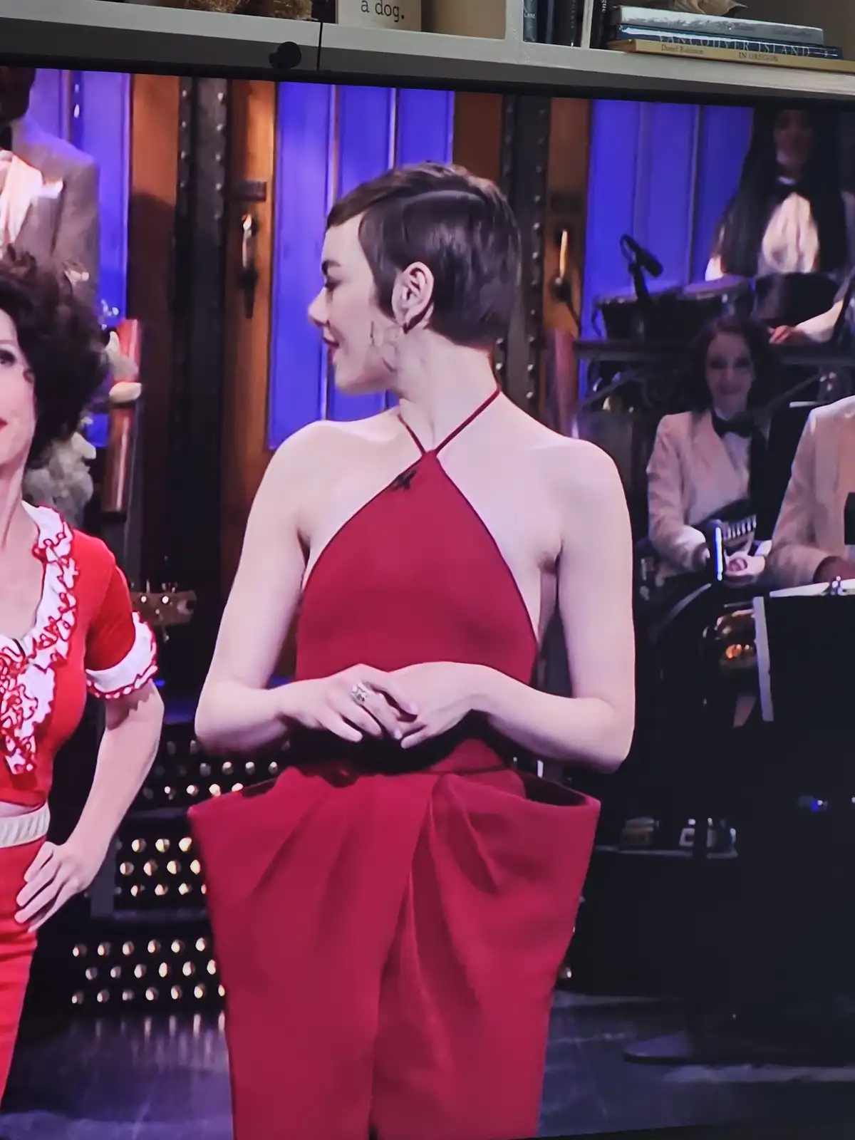 A woman with short dark hair wearing a striking red halter-neck dress with a large bow at the waist is shown in profile…