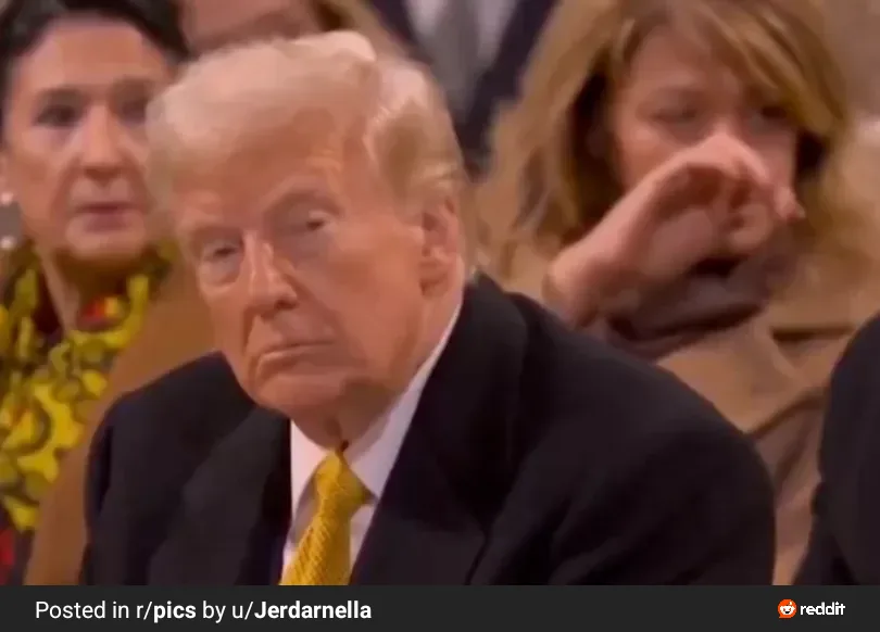 A close-up image of Donald Trump seated, looking down with a visibly displeased or bored expression, wearing a dark suit and…