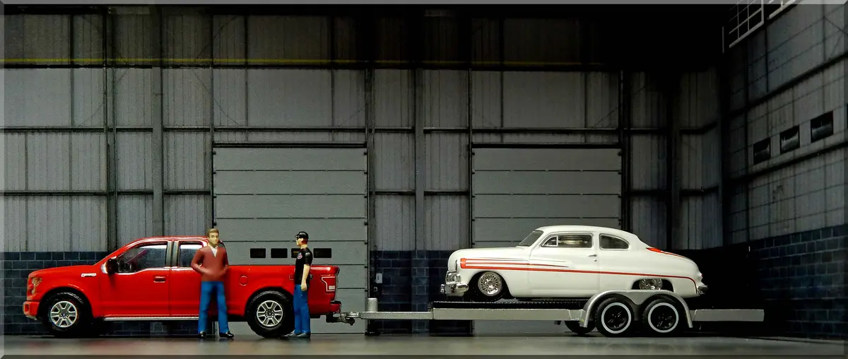 A photo of two miniature figurines standing beside a red pickup truck that is towing a trailer with a white classic car. The…