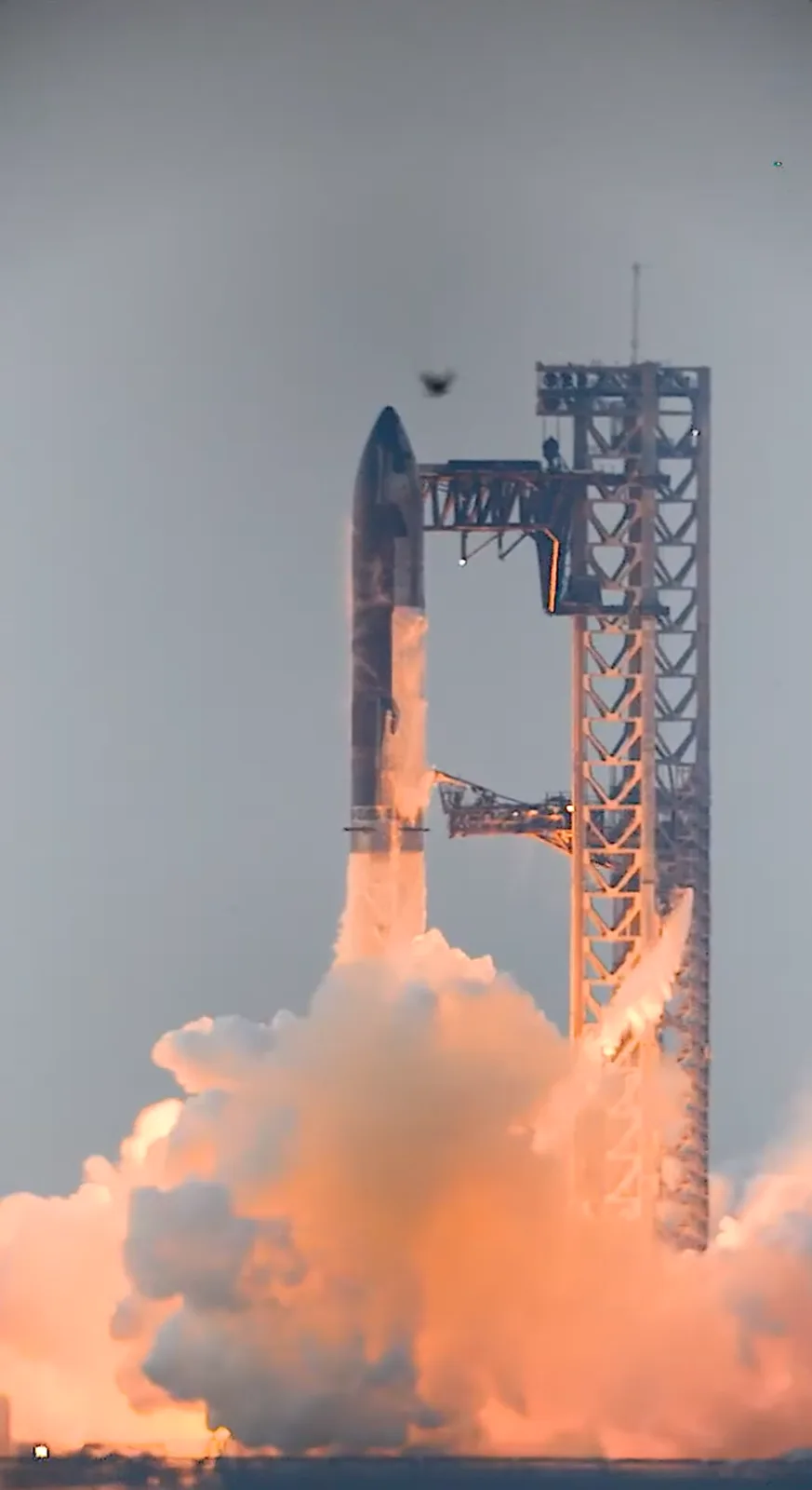 A rocket is visibly launching vertically from a launchpad, emitting a large plume of orange and white smoke and fire. A tall…