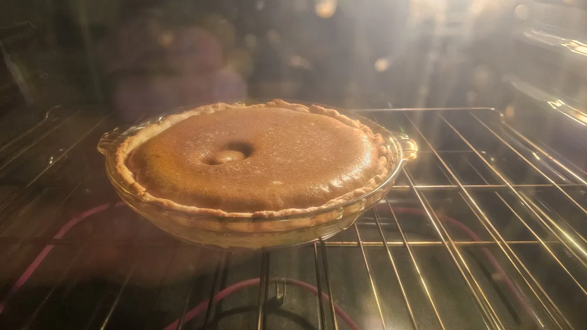 A golden-brown pie sits in a clear glass dish on an oven rack, viewed through the oven door with steam and light reflecting…