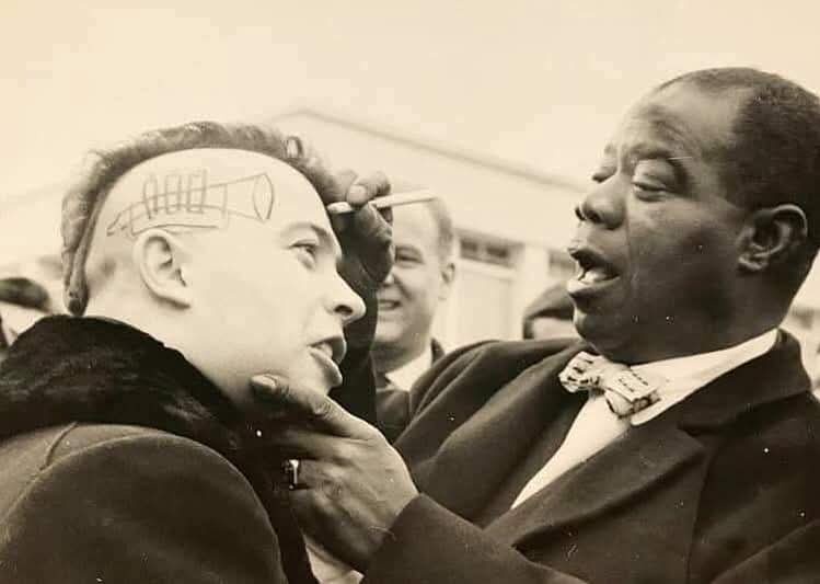 a black-and-white photo shows a man in a suit drawing a crude, cartoonish lightbulb on the shaved scalp of another man. The…