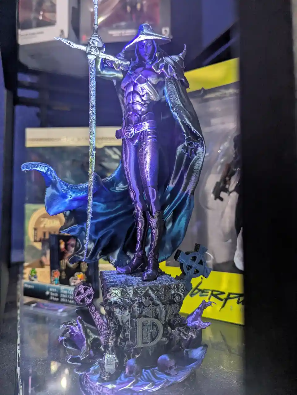A purple and blue figurine of a hooded, armored character holding a sword stands on a tombstone base with skulls and…
