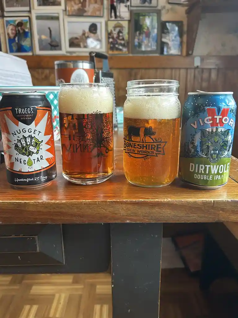 Four beer-related items sit on a wooden table: a Troegs Nugget Nectar Ale can, two mason jars filled with amber beer and…
