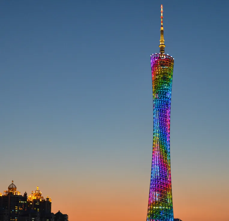 A tall, slender tower with a lattice structure is illuminated with vibrant rainbow colors against a twilight sky. The…