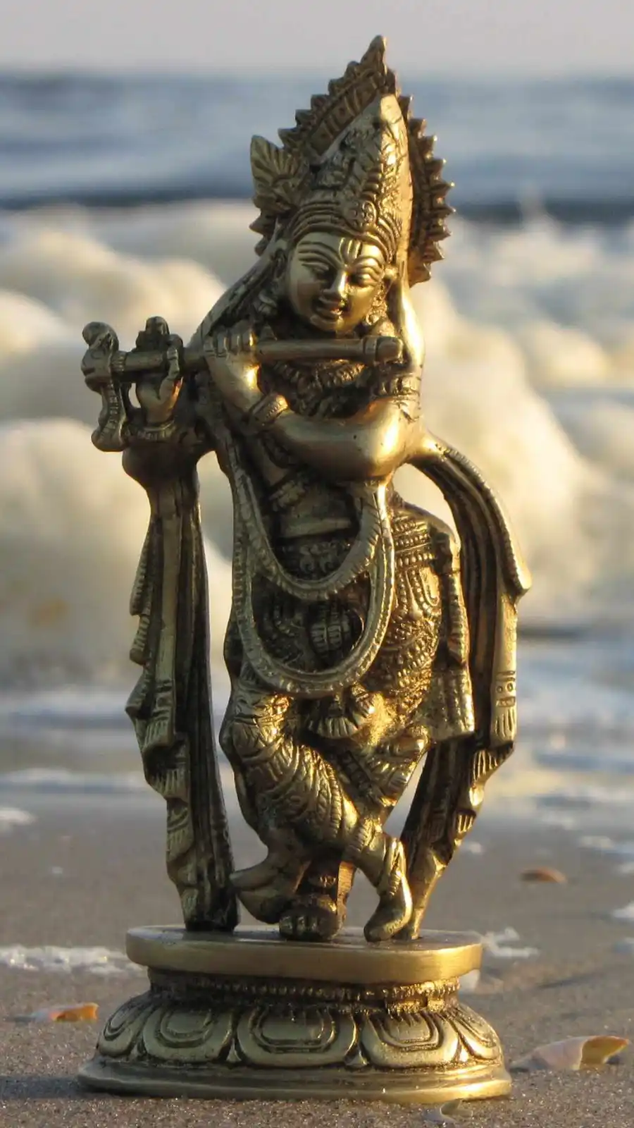 A golden statue of the Hindu deity Krishna, depicted playing a flute and dancing, is placed on a sandy beach with ocean…