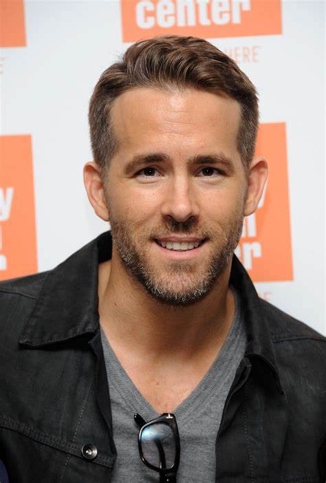 A close-up photo of actor Ryan Reynolds smiling at the camera, wearing a black jacket over a gray v-neck shirt with glasses…