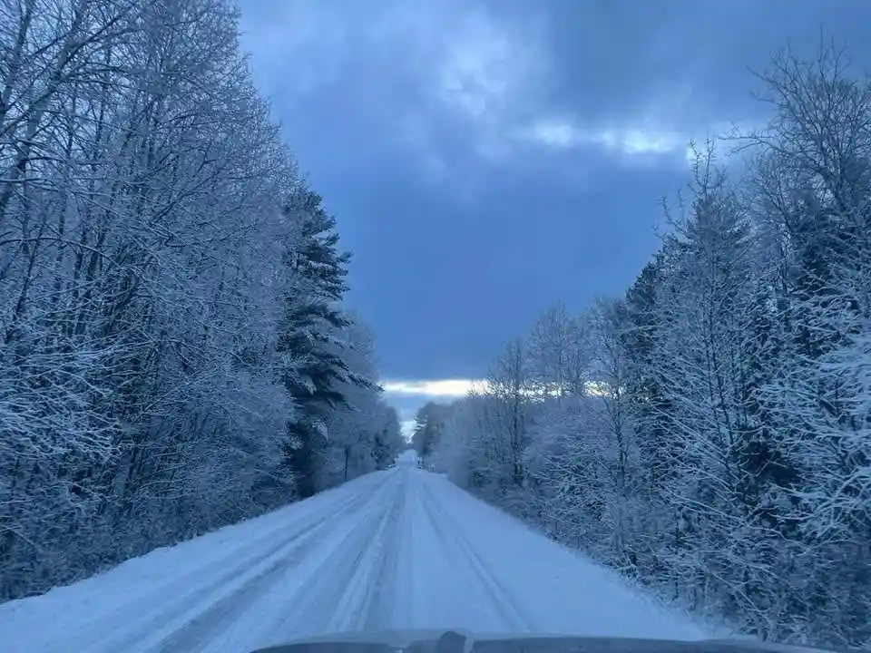 A point-of-view shot from inside a car driving down a snow-covered road flanked by snow-laden trees under a cloudy blue sky…