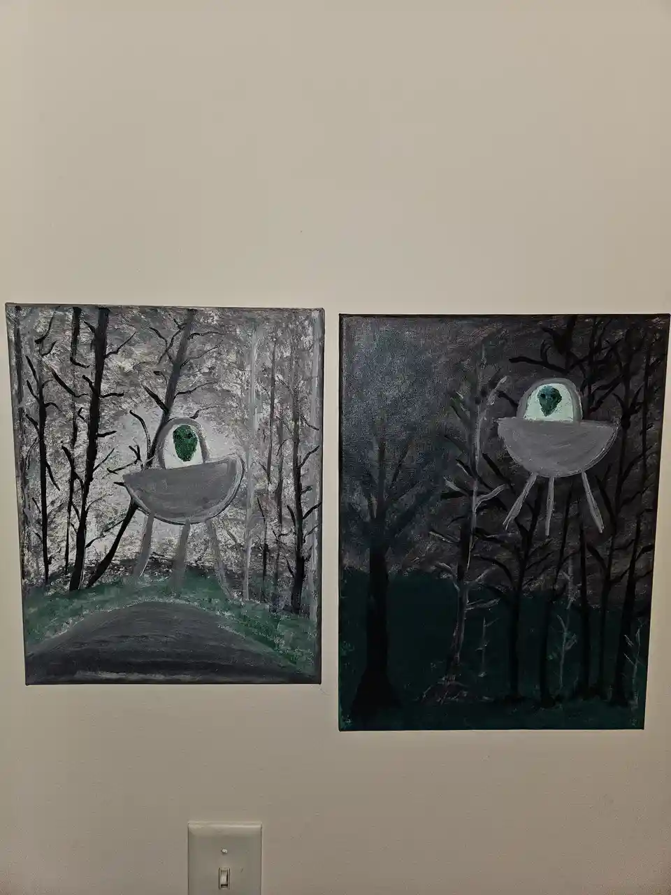 Two hand-painted canvases hang side-by-side on a wall, each depicting a gray, bowl-shaped alien with a green face peeking…