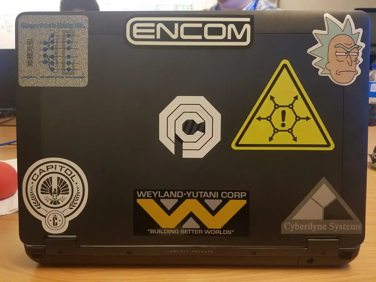 The back of a black laptop covered in stickers from fictional sci-fi franchises, including ENCOM, Weyland-Yutani Corp…