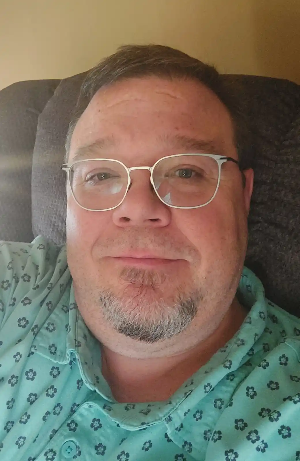 A man with glasses and a goatee, wearing a light green floral shirt, is reclining in a dark gray chair. He has a subtle…