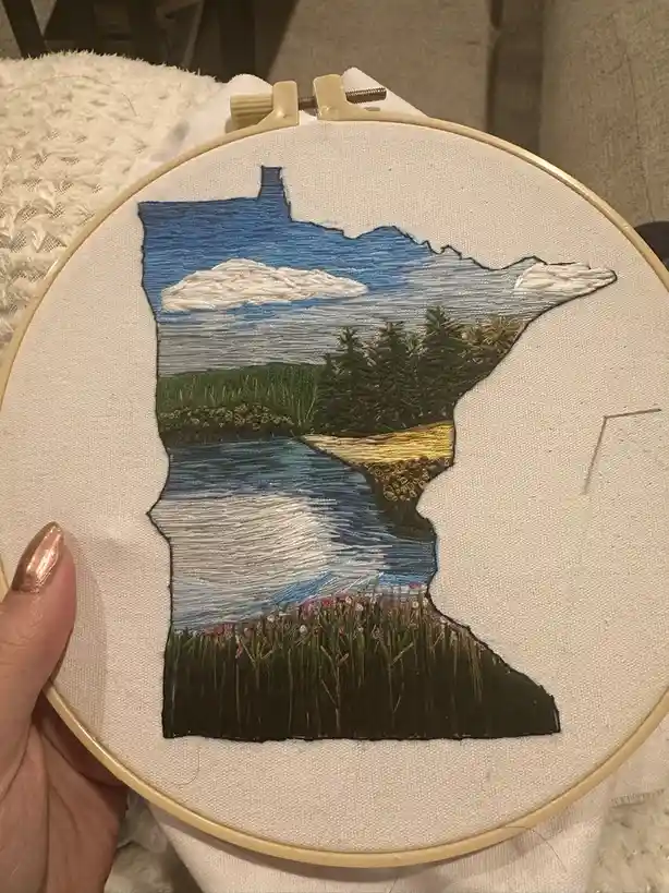 A hand holds an embroidery hoop displaying a detailed stitched artwork shaped like the state of Minnesota. The design within…
