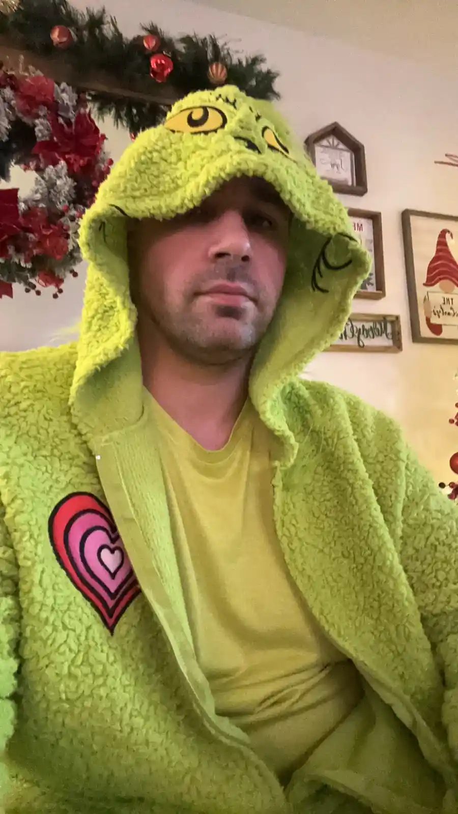 A man wearing a bright green, fuzzy Grinch-themed hoodie with a large cartoon eye on the hood, looks directly at the camera…