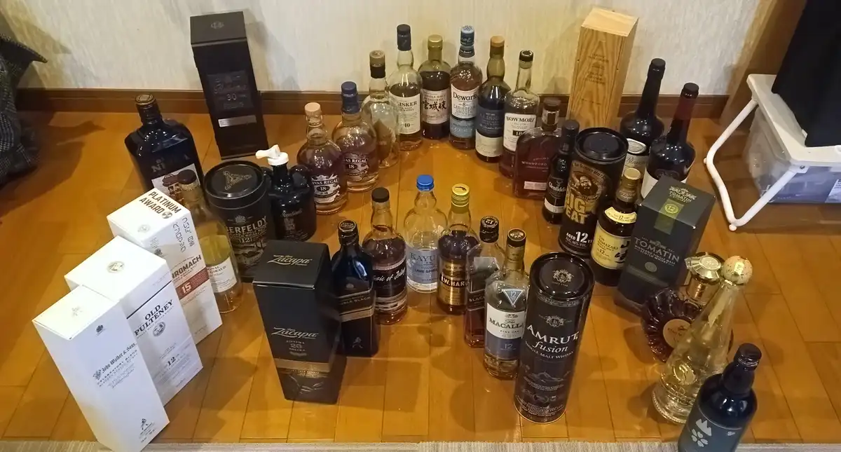 An overhead photo shows a large collection of whiskey bottles and boxes arranged on a wooden floor, including brands like…