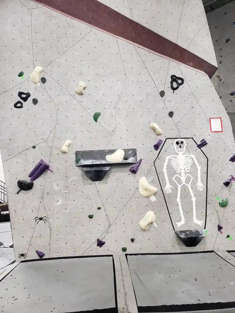An indoor climbing wall features a large white outline of a skeleton inside a coffin drawn on its surface, surrounded by…