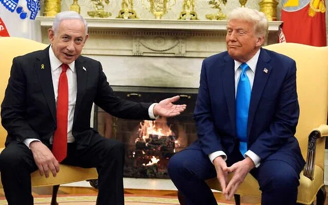 two men sit in ornate chairs in front of a fireplace; the man on the left gestures with his hand while looking at the man on…