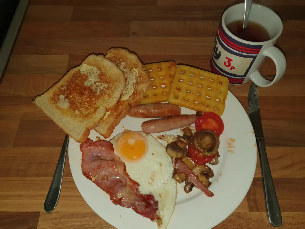 A plate of a full English breakfast including fried egg, bacon, sausages, mushrooms, tomatoes, toast with butter, and waffle…